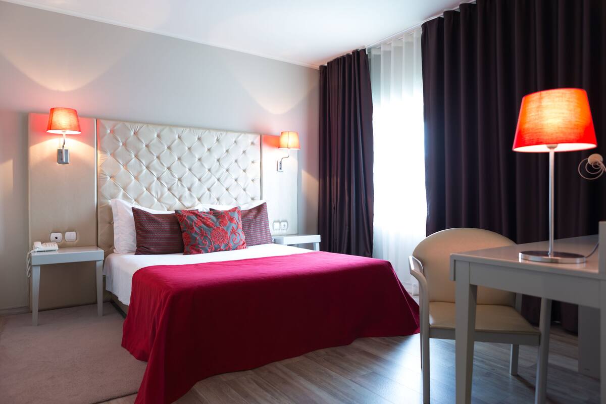 Een hotelkamer met een gestoffeerd hoofdeinde, een rode sprei, twee bedlampen, een bureau en een stoel.