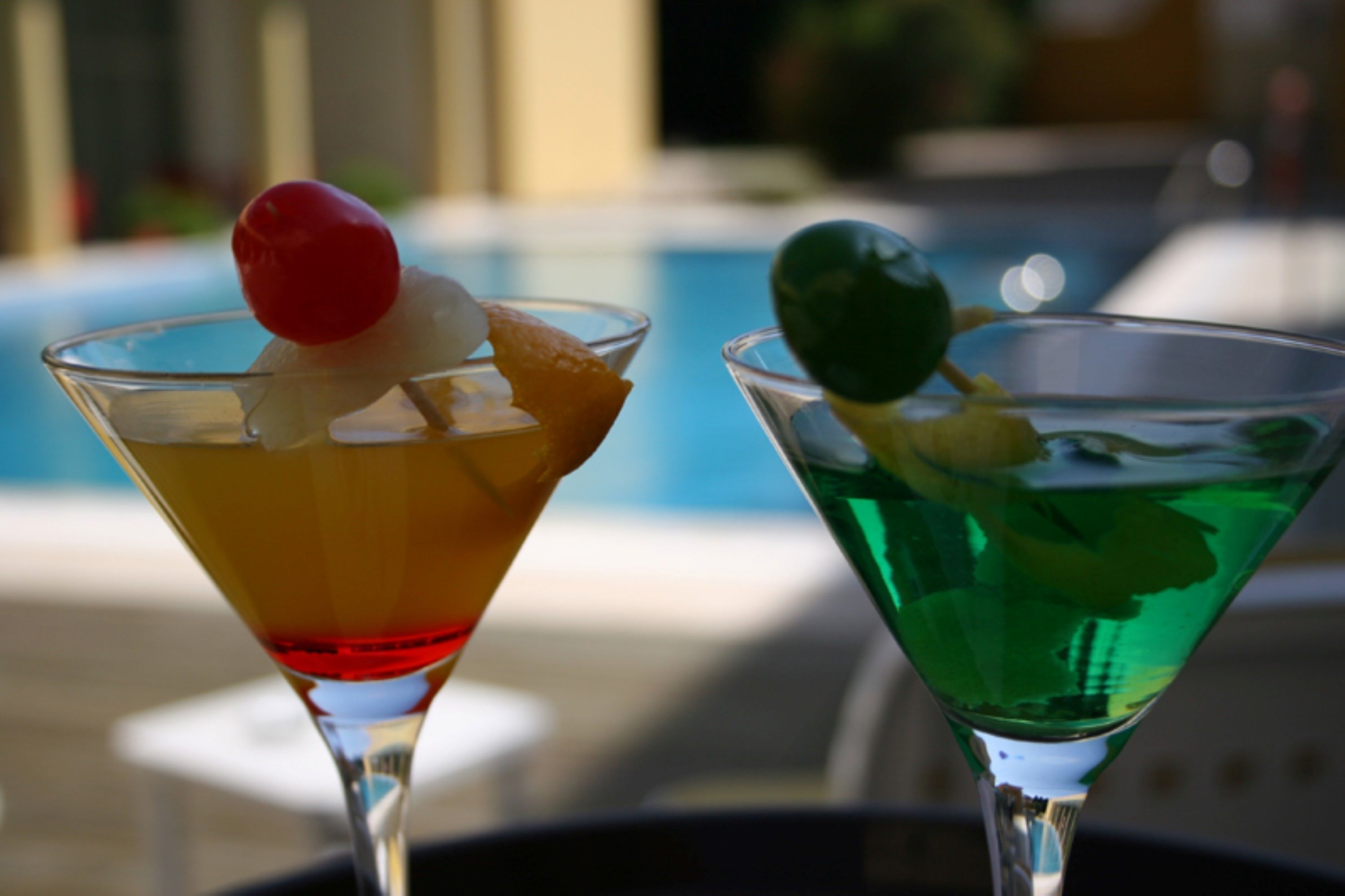 Two cocktails on a table near a pool, with a blurred background suggesting an outdoor setting.