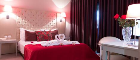 A hotel room with a tufted headboard, red bedding, a vase with red roses, and a white table with a lamp.