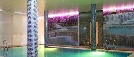 An indoor swimming pool with a waterfall feature, surrounded by walls with a mosaic tile design.