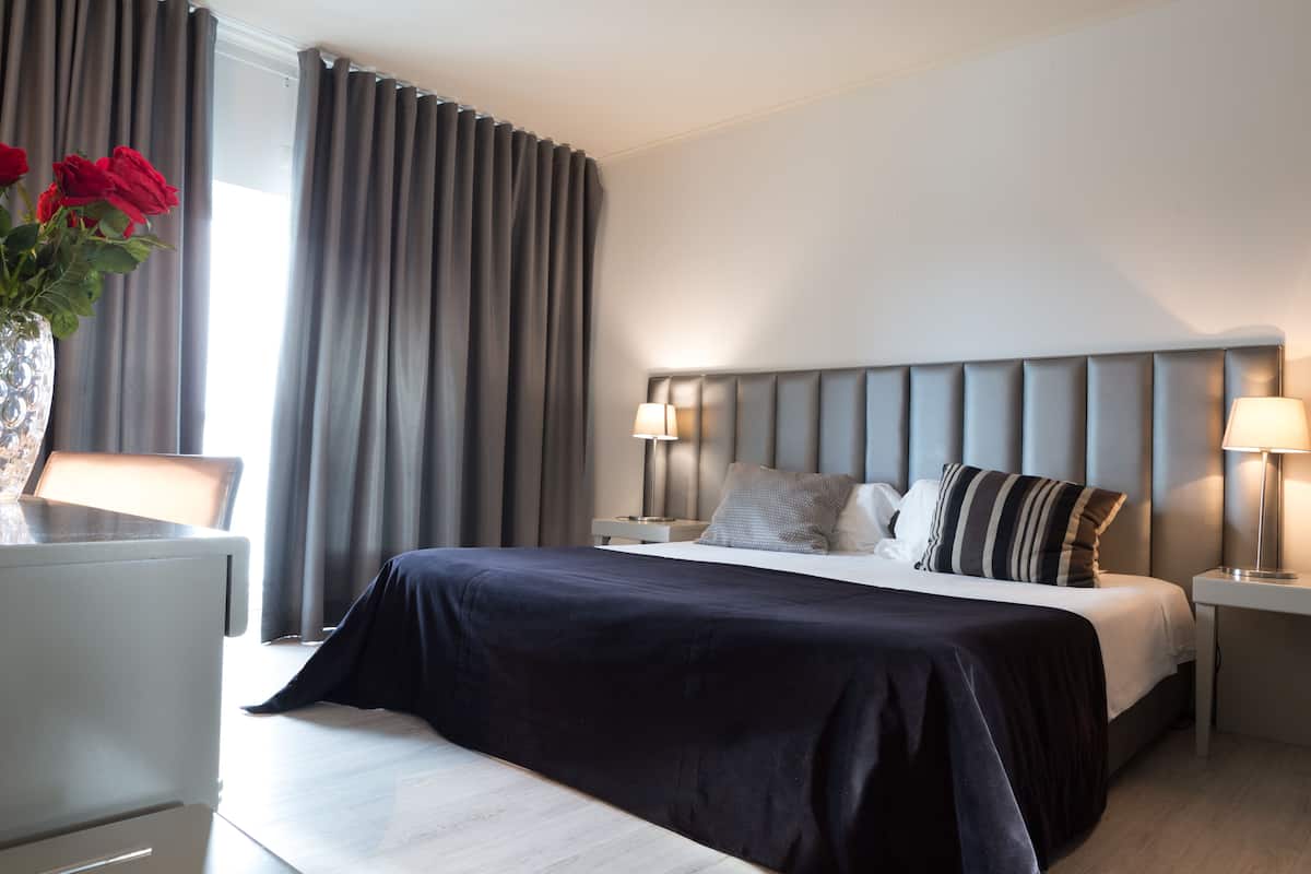 Een moderne hotelkamer met een groot bed, twee bedlampjes, een nachtkastje en een vaas met rode bloemen.