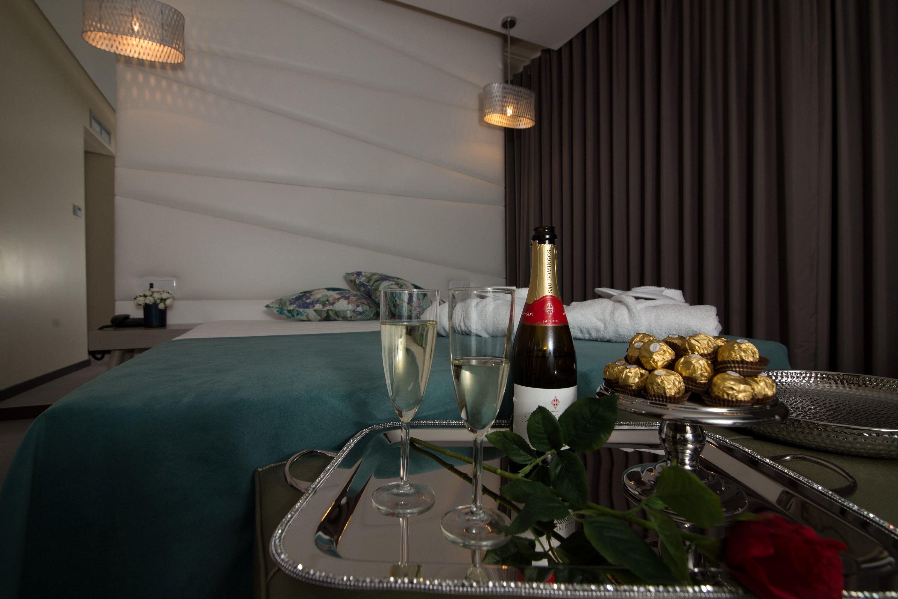 A neatly arranged hotel room with a bed, a bedside table with a tray of champagne glasses, a bottle of champagne, and chocolates.