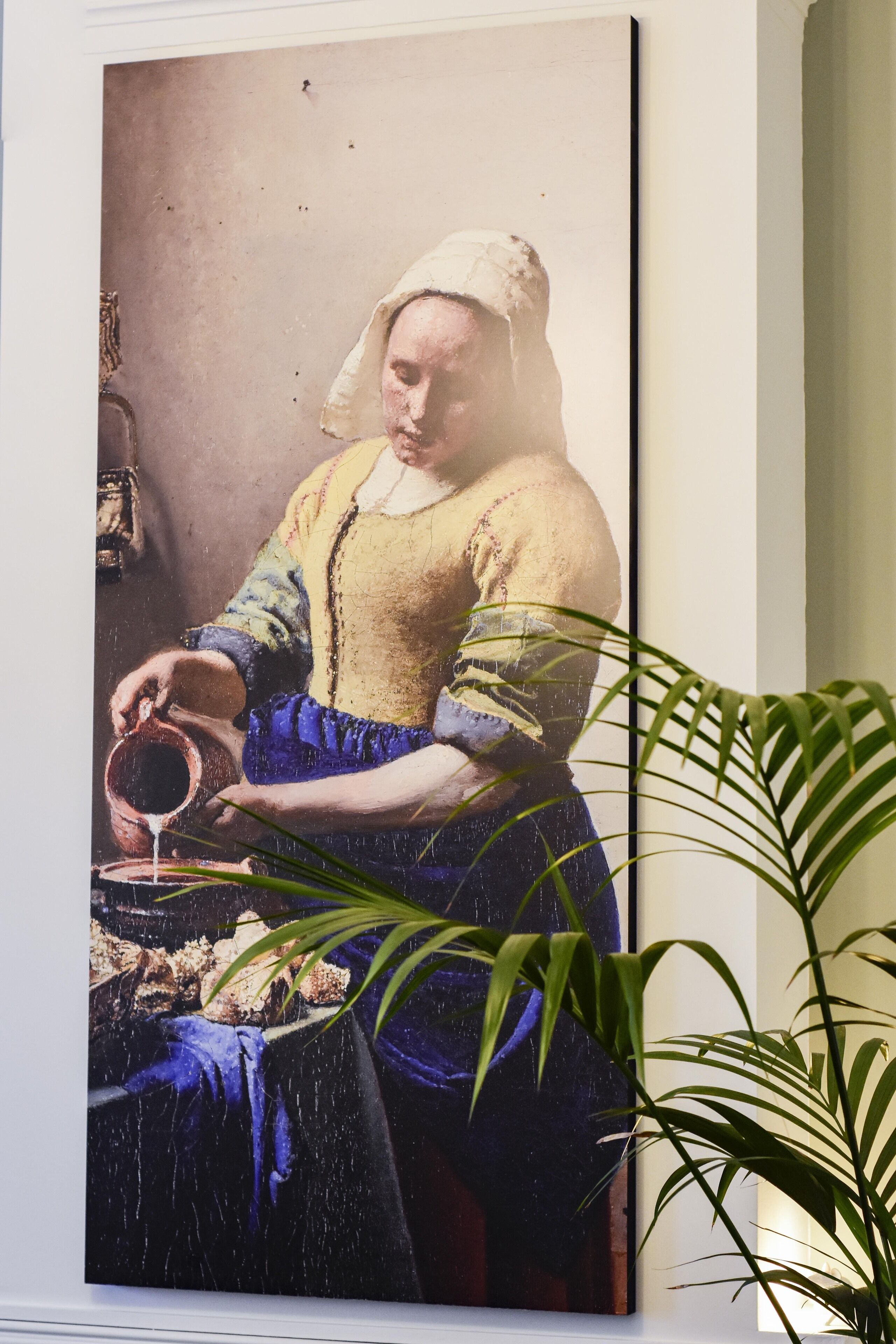 A framed painting of a person pouring liquid, with a potted plant in the foreground.