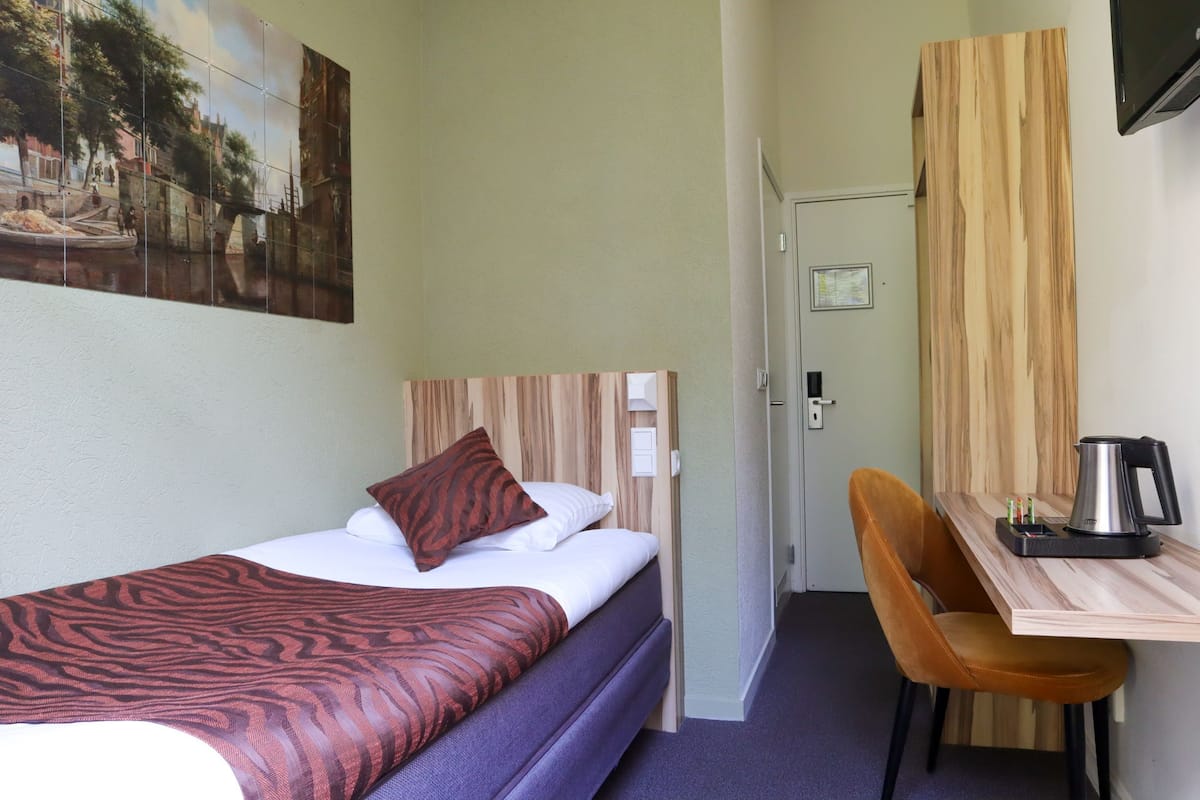 A single bed with a zebra-patterned blanket, a wooden headboard, and a brown pillow. There is a wooden desk with a chair and a kettle on it.