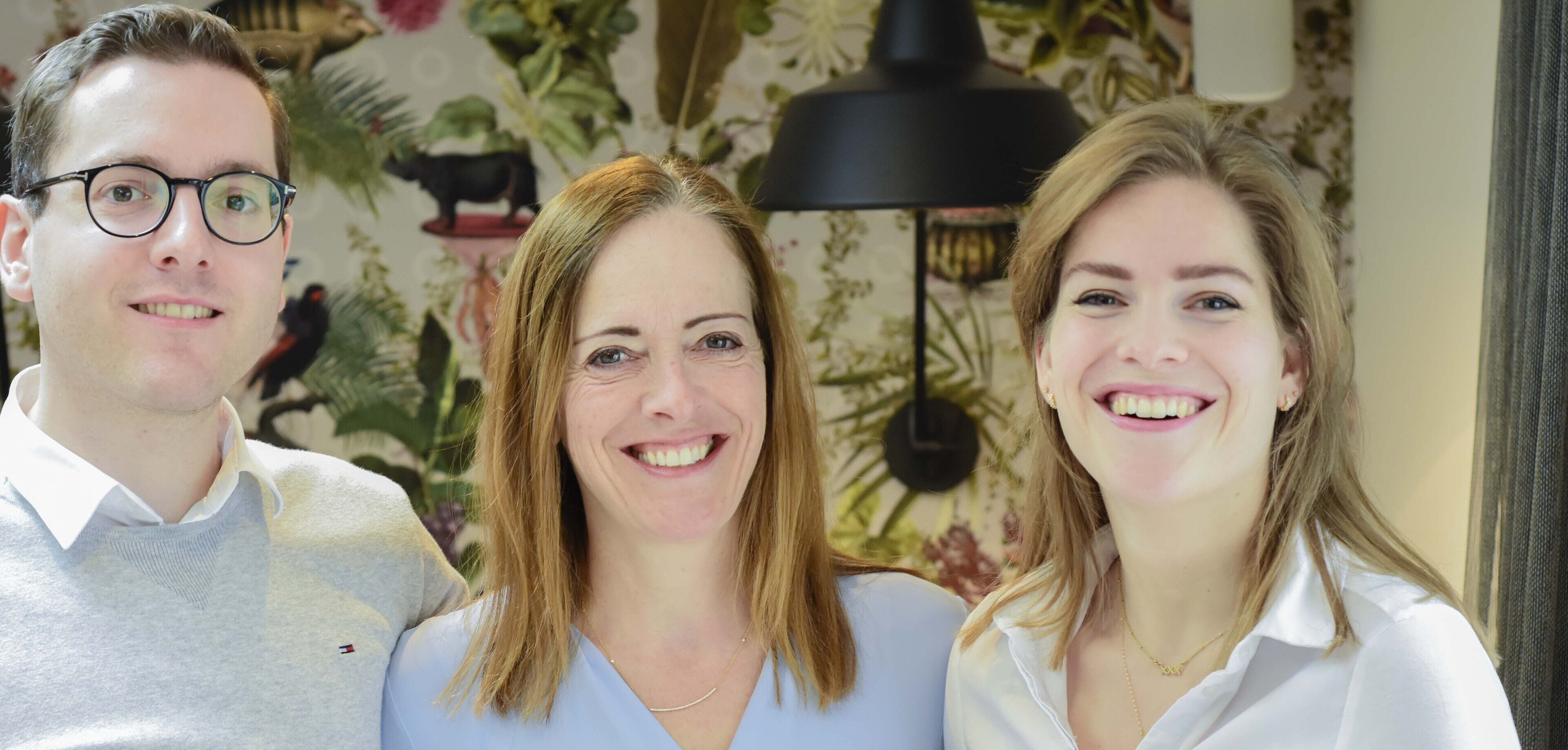 Three people standing in a room with floral wallpaper and a black lamp.