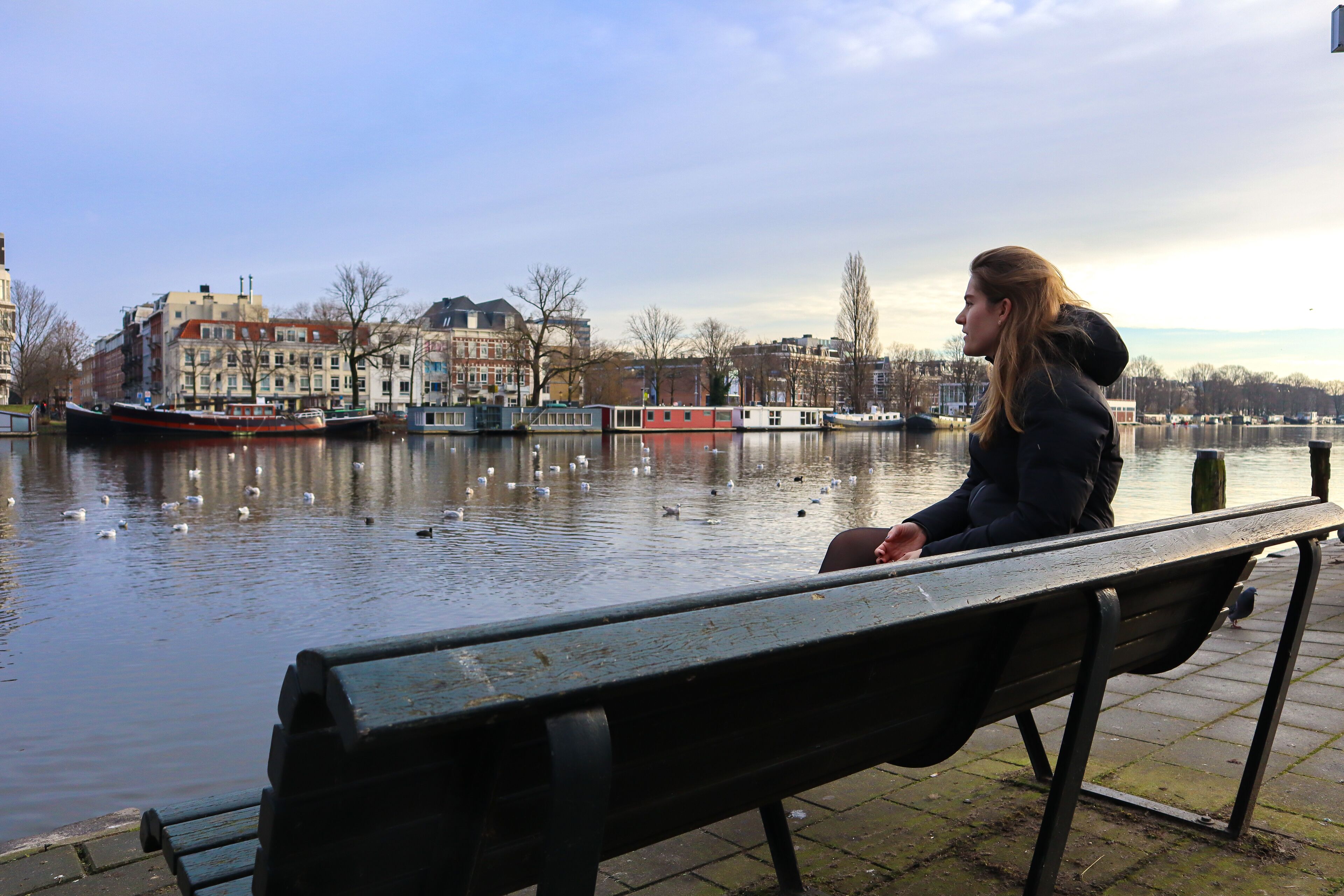 A person sitting on a bench by a calm river with buildings in the background.