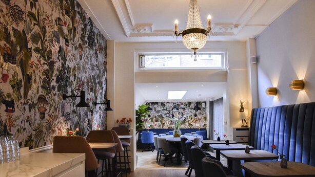 A restaurant with a high ceiling, a chandelier, and a floral wallpaper design. There are multiple tables and chairs, and a blue booth area. - Boutique Hotel Asterisk, a family run hotel (Amsterdam)