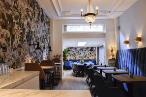 Ein Restaurant mit hohen Decken, einem Kronleuchter und einem Wanddesign mit floralem Muster. Es gibt mehrere Tische und Stühle sowie eine Sitzgruppe als Box.