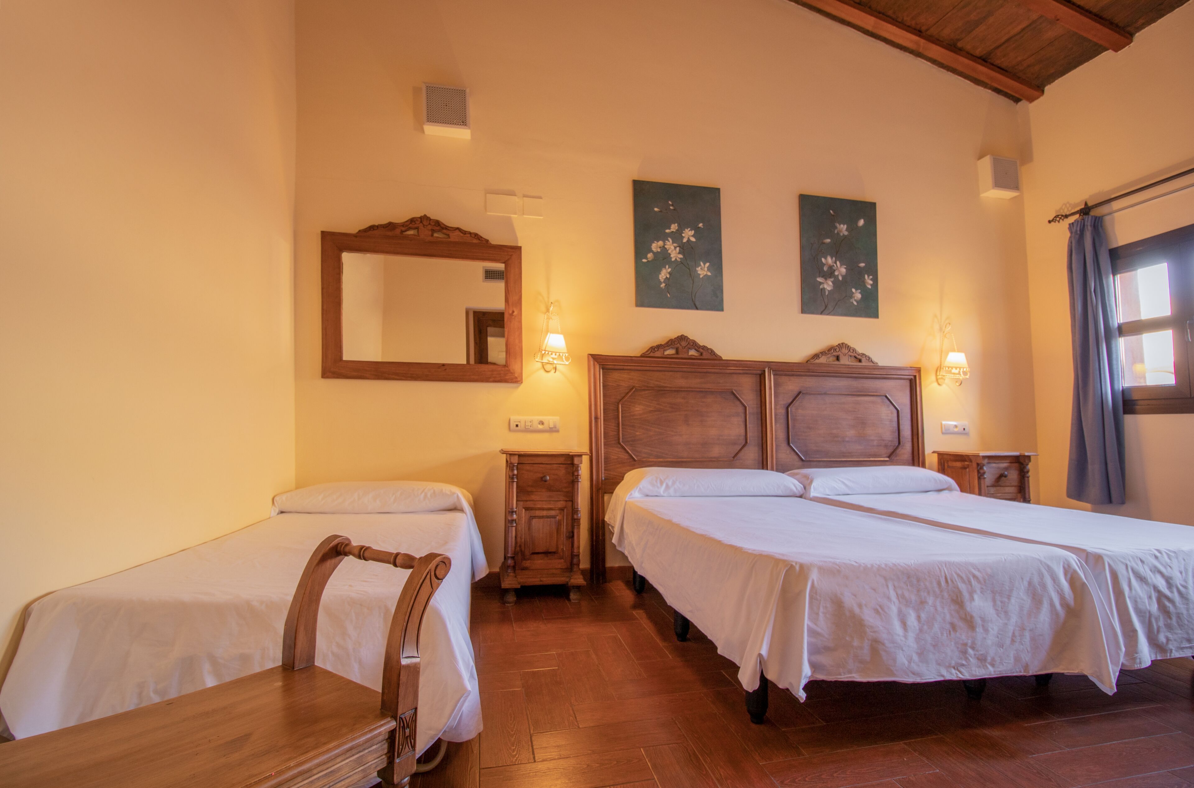 A room with two single beds, wooden headboards, a mirror, wall-mounted lights, and a window with curtains.
