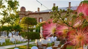 An outdoor dining area with round tables and white chairs, surrounded by greenery and a stone building in the background.