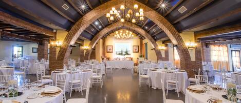 A spacious banquet hall with white tables and chairs, a large chandelier, and brick arches.