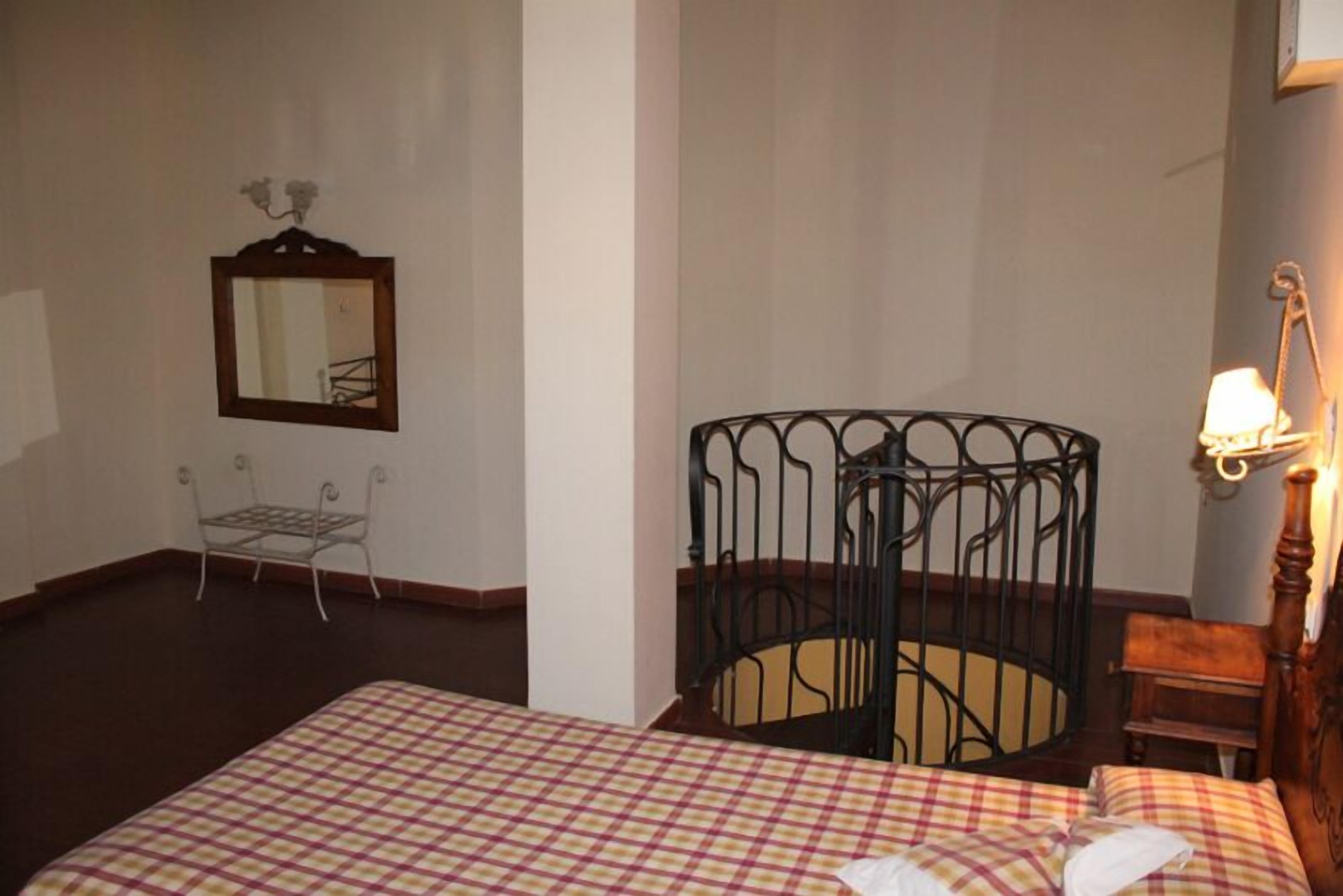 Una habitación de hotel con una colcha a cuadros, una silla de estructura metálica, una mesita pequeña, un cabecero de cama metálico y un espejo fijado a la pared.