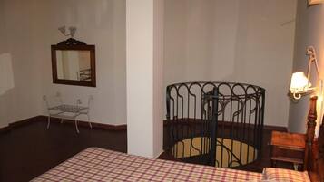 Una habitación de hotel con una colcha a cuadros, una silla de estructura metálica, una mesita pequeña, un cabecero de cama metálico y un espejo fijado a la pared.