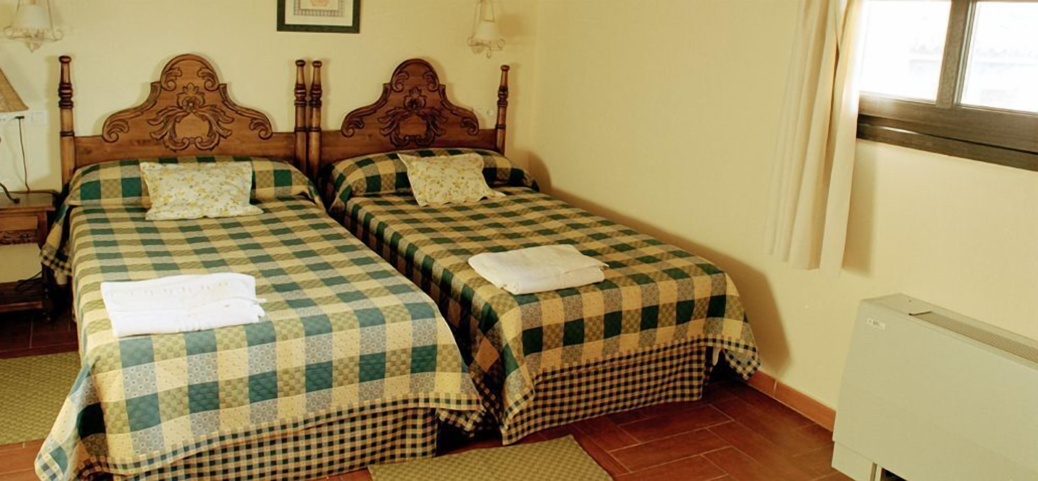 Habitación con dos camas, cada una con una colcha a cuadros y una almohada. Hay una mesita de noche con una lámpara, un radiador y una ventana con cortinas.