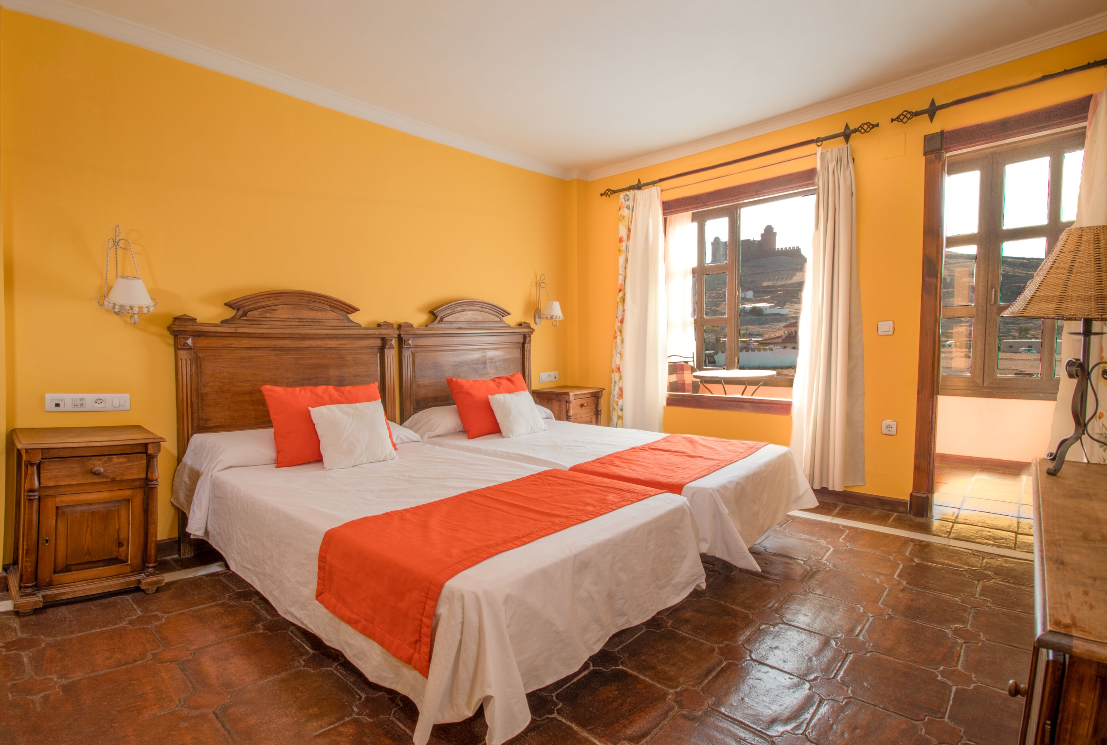 Una cama doble con ropa de cama blanca y naranja, una mesita de noche de madera y vistas a un balcón con barandilla.