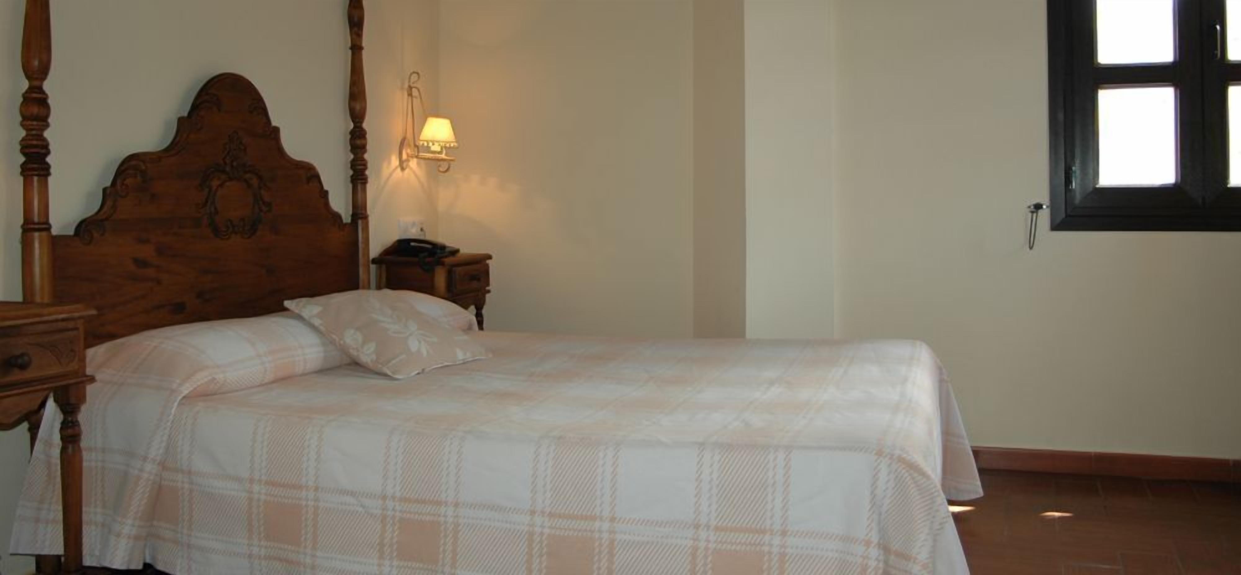A bedroom with a wooden four-poster bed, a bedside table with a lamp, and a window with a view.