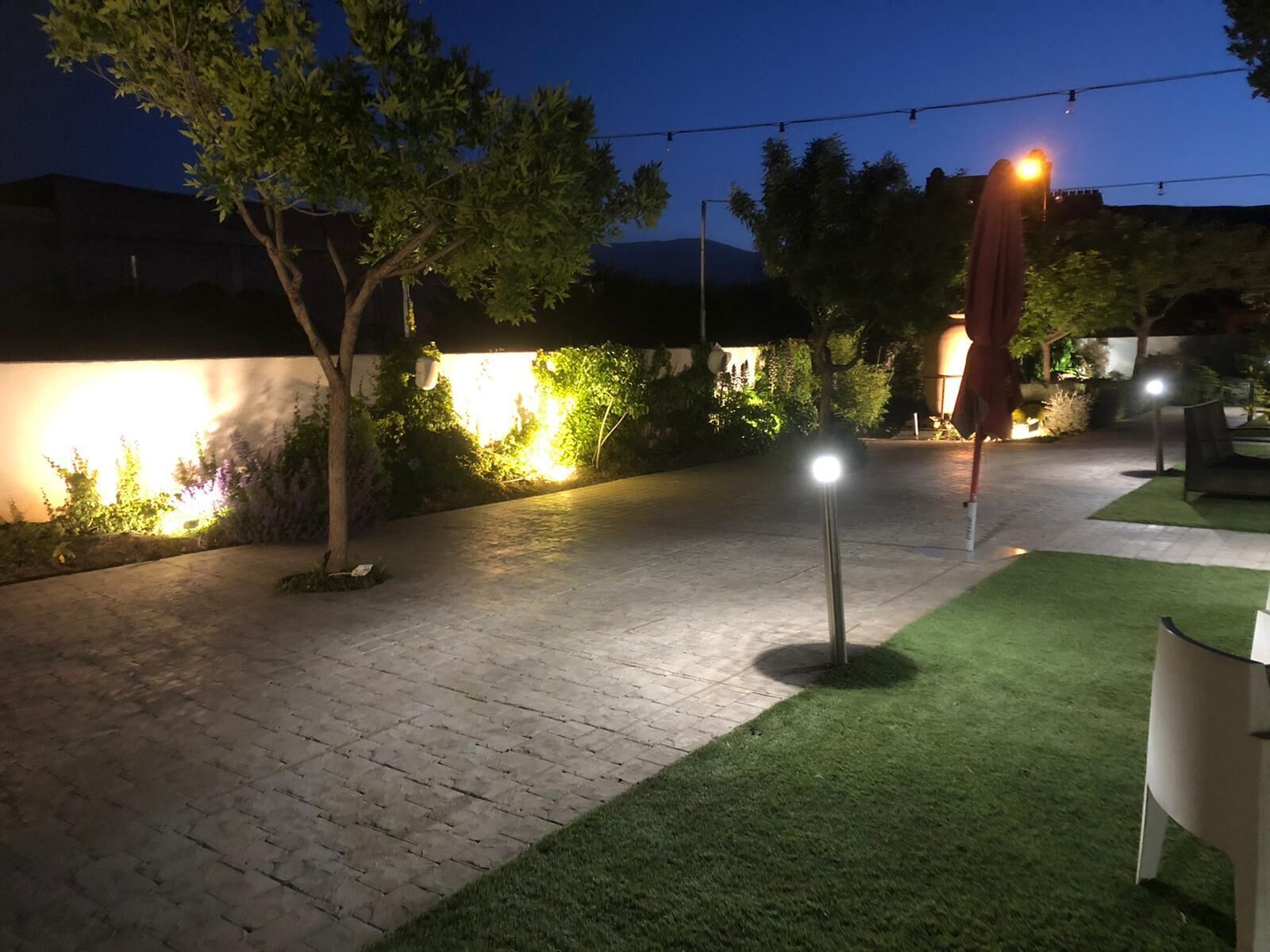 Un patio al aire libre con un camino empedrado, césped artificial verde y luces colgantes.