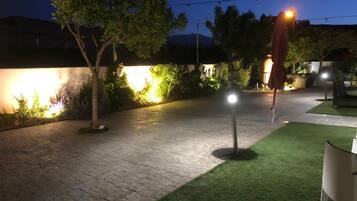 Un patio al aire libre con un camino empedrado, césped artificial verde y luces colgantes.
