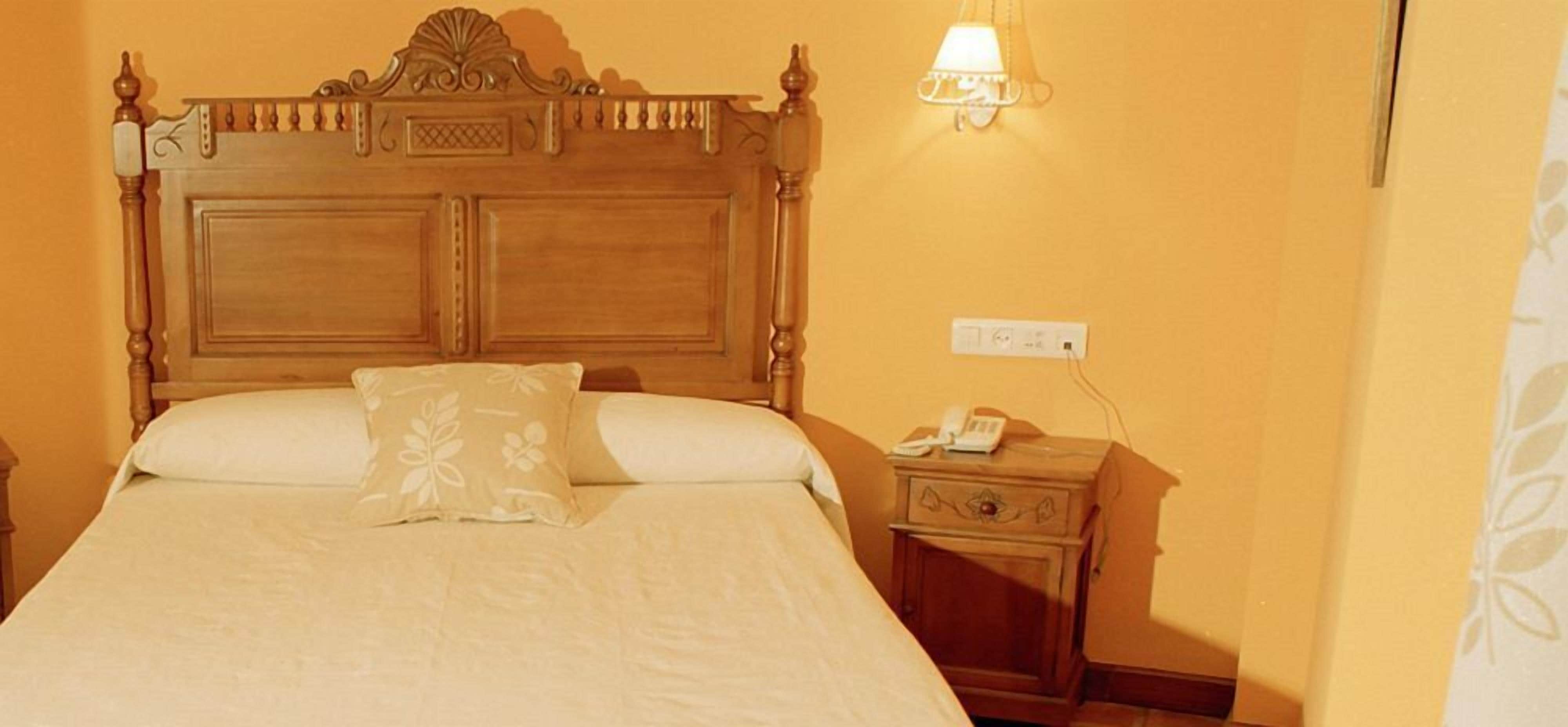 Una cama bien hecha con cabecera de madera, una mesita de noche con lámpara y un enchufe en la pared.