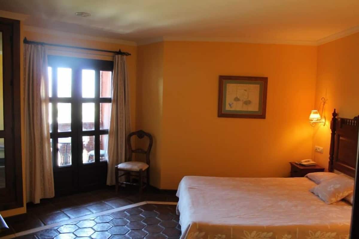A hotel room with a bed, a chair, a window with curtains, a framed picture on the wall, and a nightstand with a lamp.