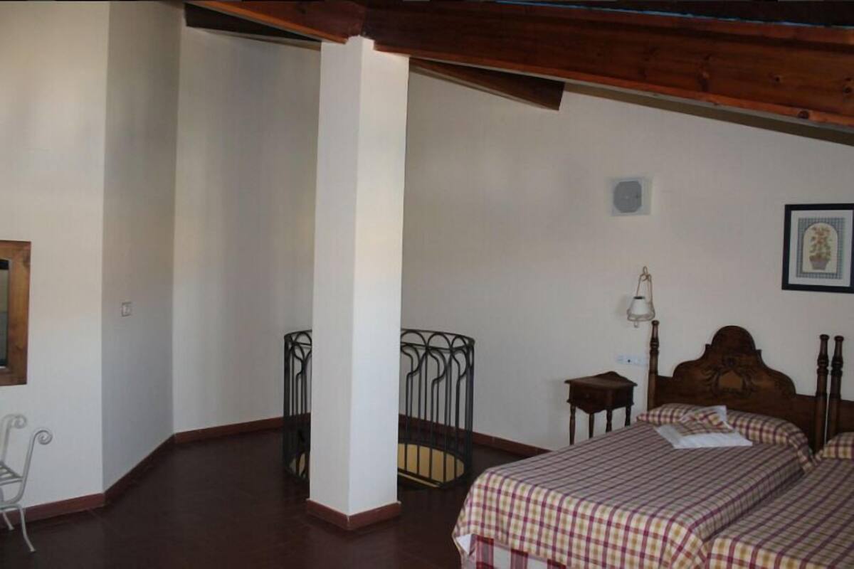 A bedroom with a wooden ceiling, a bed with a checkered bedspread, a nightstand, a chair, and a small table.