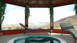 A circular hot tub with two lounge chairs, surrounded by a red wall and large windows with greenery.