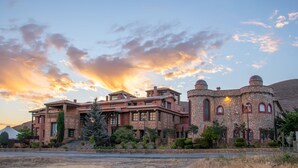 A large, historic building with multiple levels and a stone facade, surrounded by landscaped gardens and a clear sky.