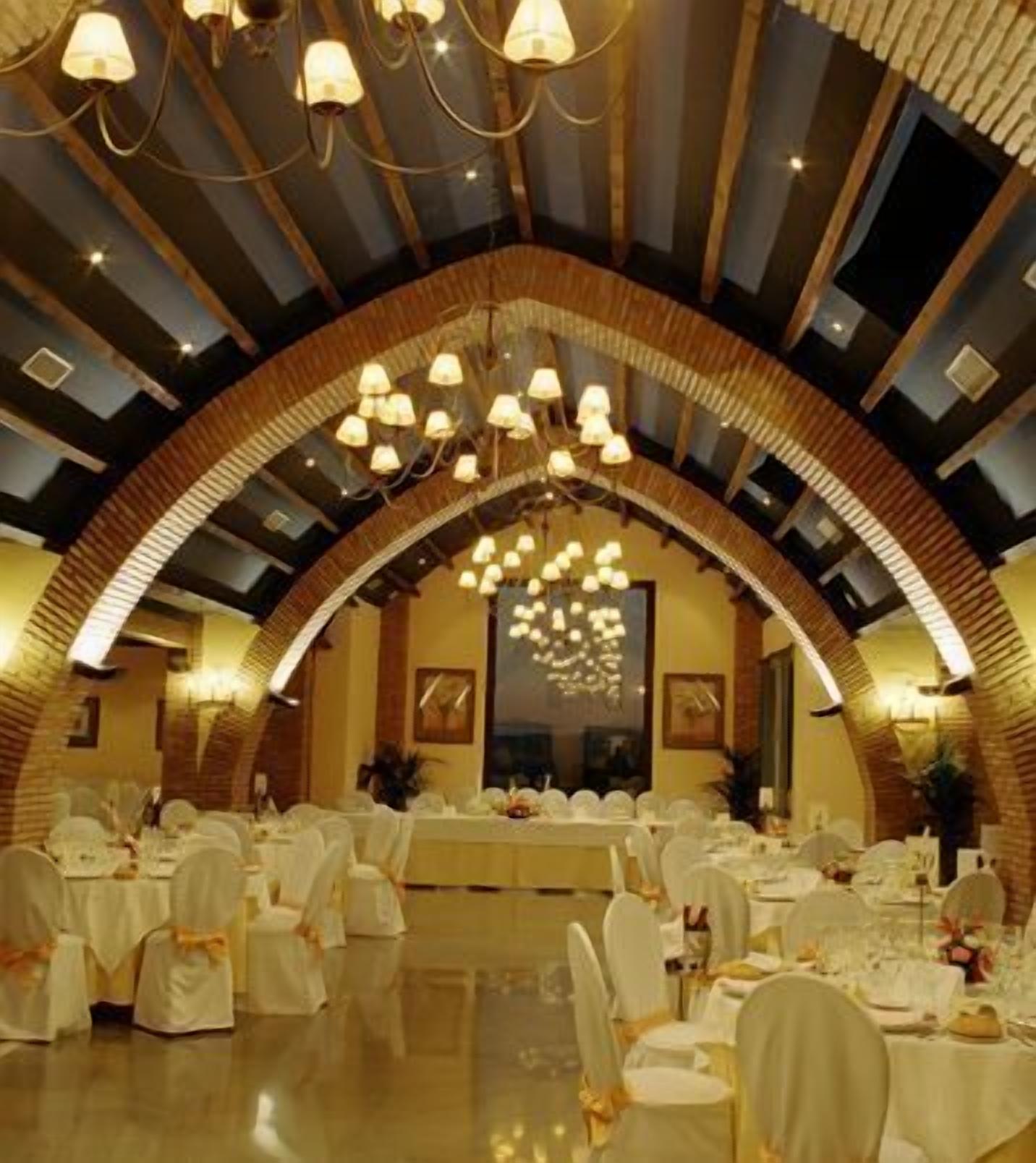 A banquet hall with round tables and chairs, a high ceiling with chandeliers, and brick arches.