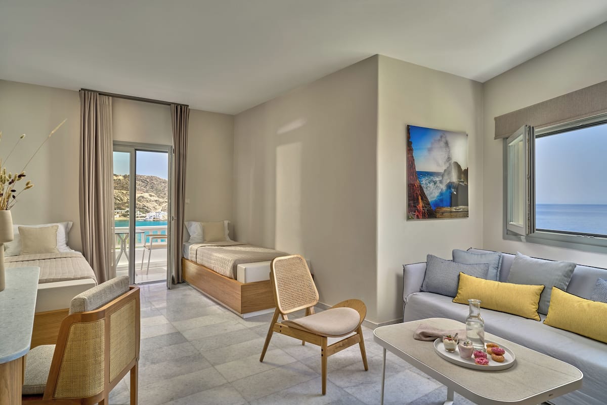 A hotel room with a bed, a sofa, a dining table, and a chair. There is a window with a view of the sea.