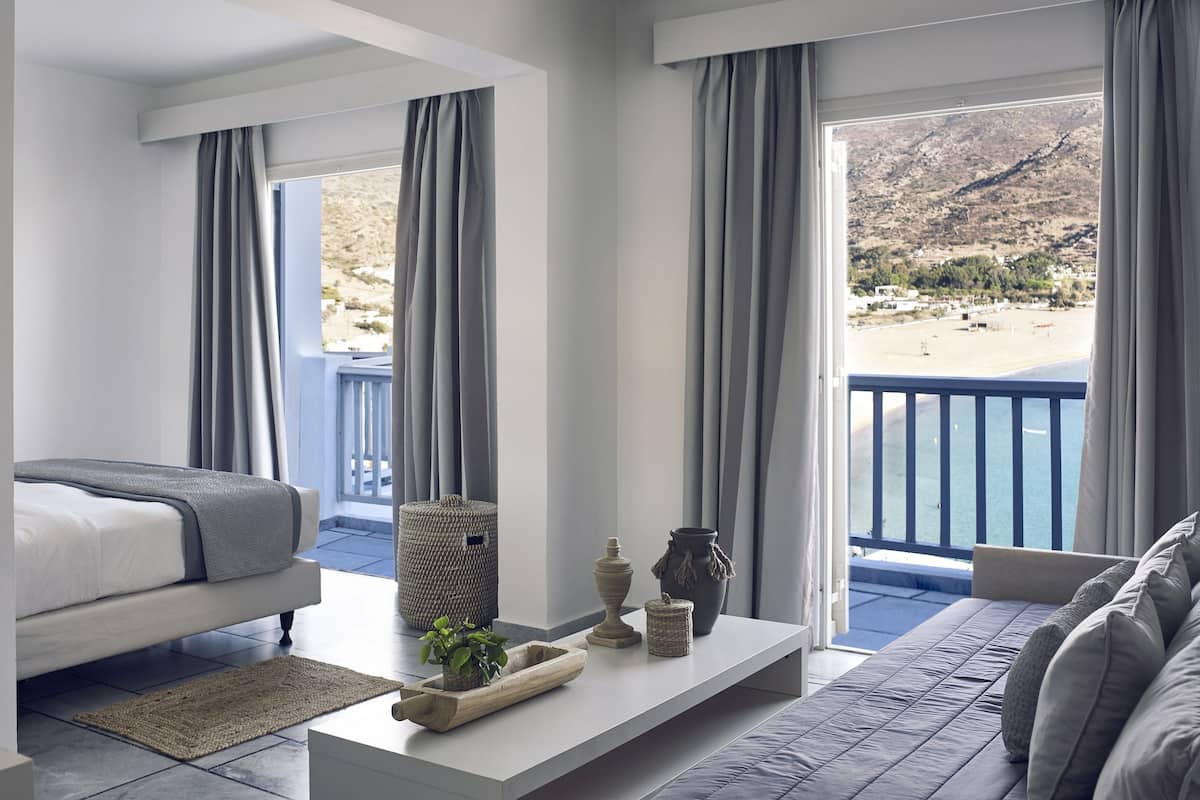 A modern hotel room with a bed, a sofa, a small table with decorative items, and a view of a beach and mountains.