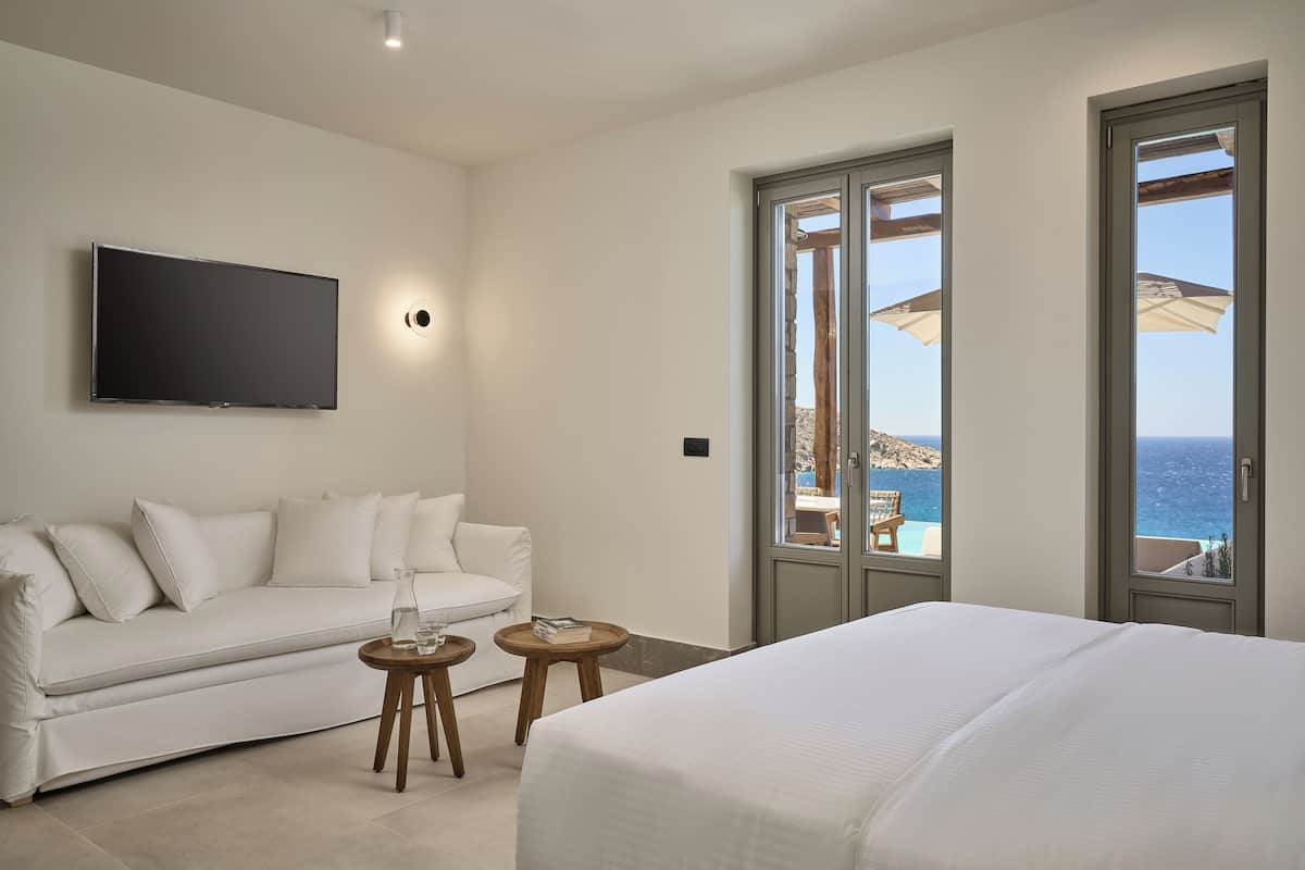 A modern hotel room with a white sofa, a bed, a TV, and a balcony with a view of the sea.