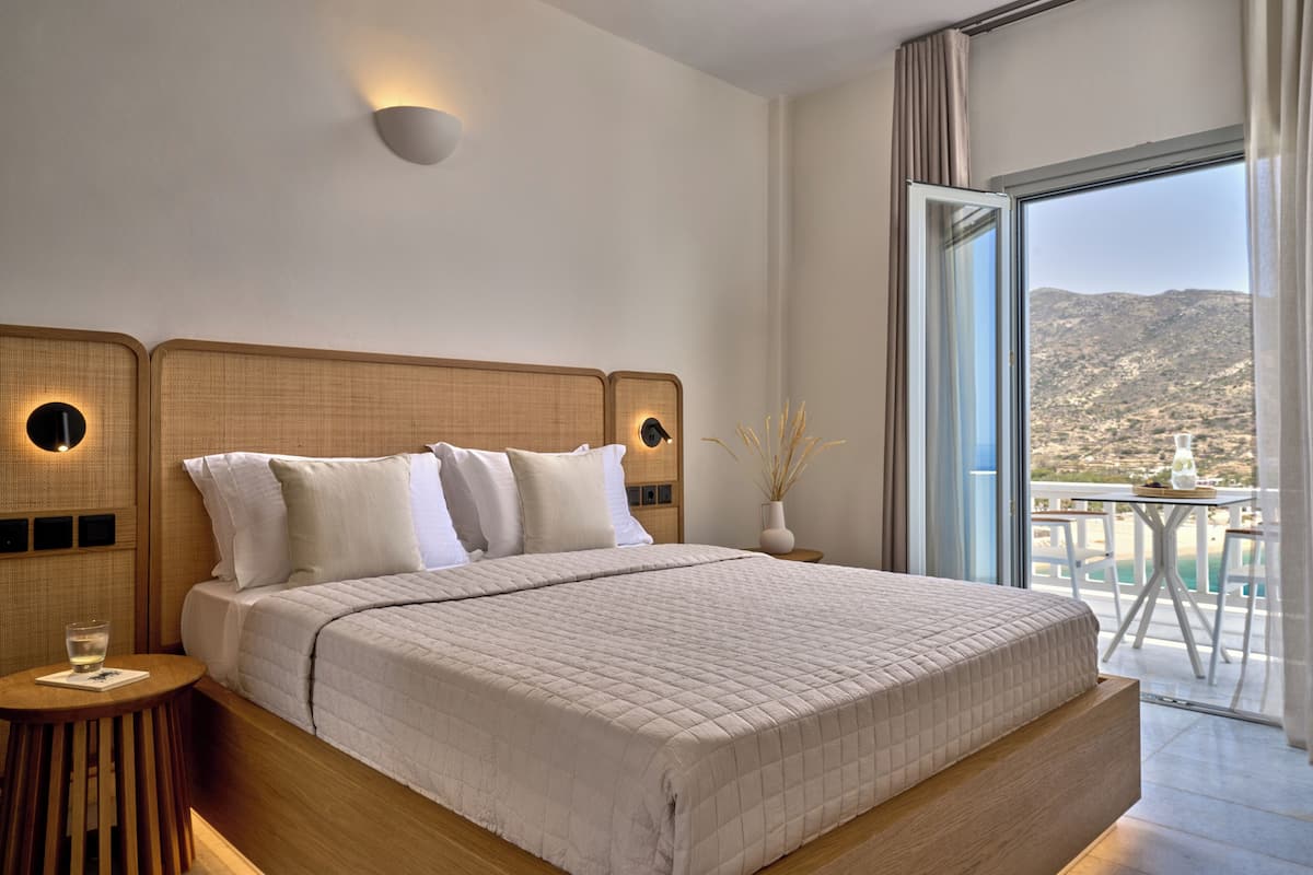 A modern bedroom with a large bed, a bedside table with a glass, and a view of a balcony with a table and chairs.