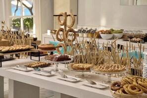 Uma mesa de buffet com diversos produtos de panificação, incluindo tortas, biscoitos e doces, dispostos em travessas brancas.