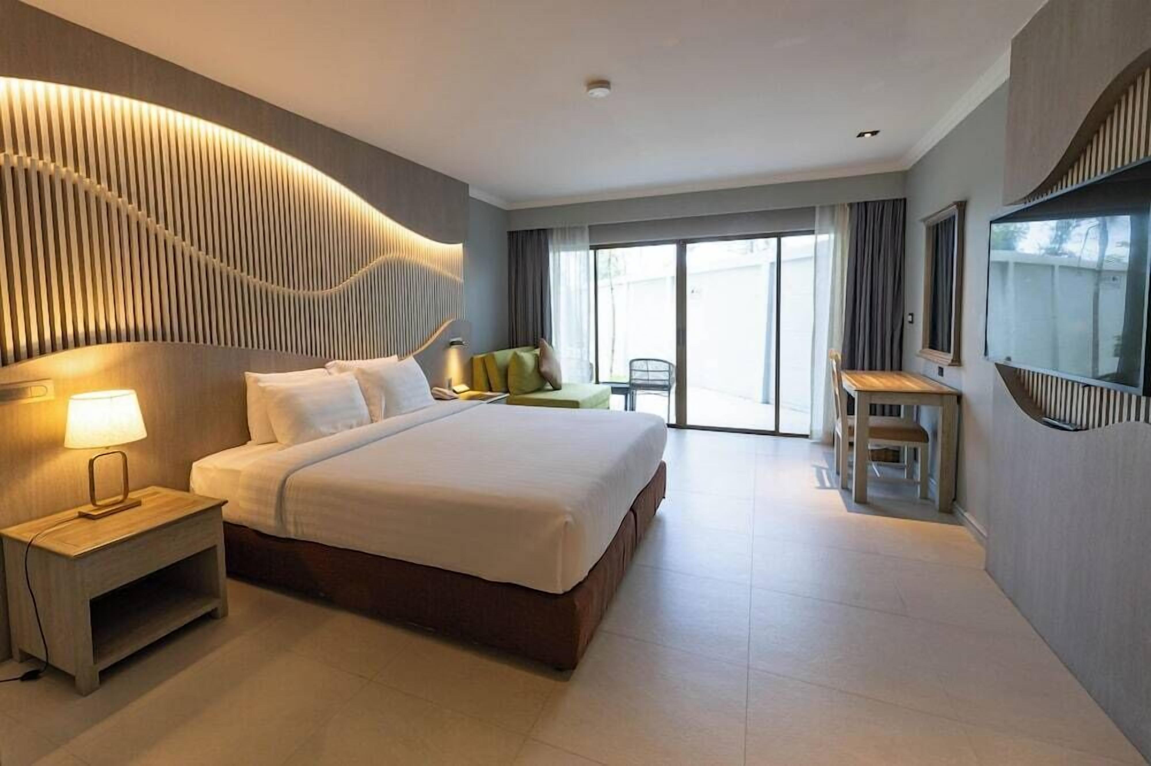 A modern hotel room with a large bed, a bedside table with a lamp, a desk with a chair, and a large window with curtains.