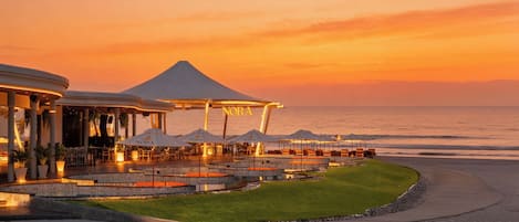 A waterfront restaurant with outdoor seating, a covered area, and a view of the ocean at sunset.