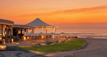 Ein Restaurant am Wasser mit Terrassensitzgelegenheiten, einem überdachten Bereich und Blick auf den Sonnenuntergang über dem Meer.
