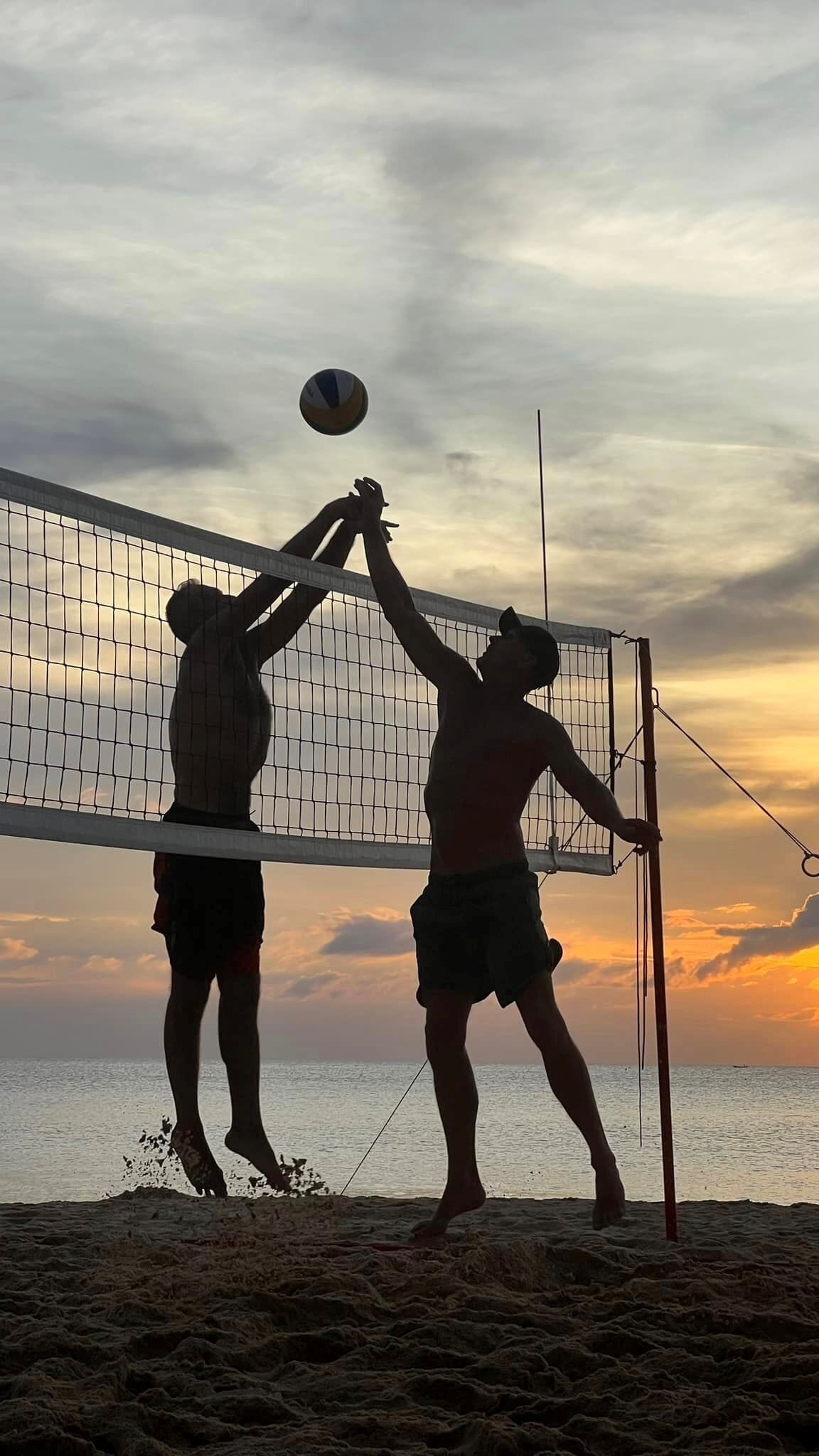 Two people playing beach volleyball on a sandy court with a net, under a sunset sky.