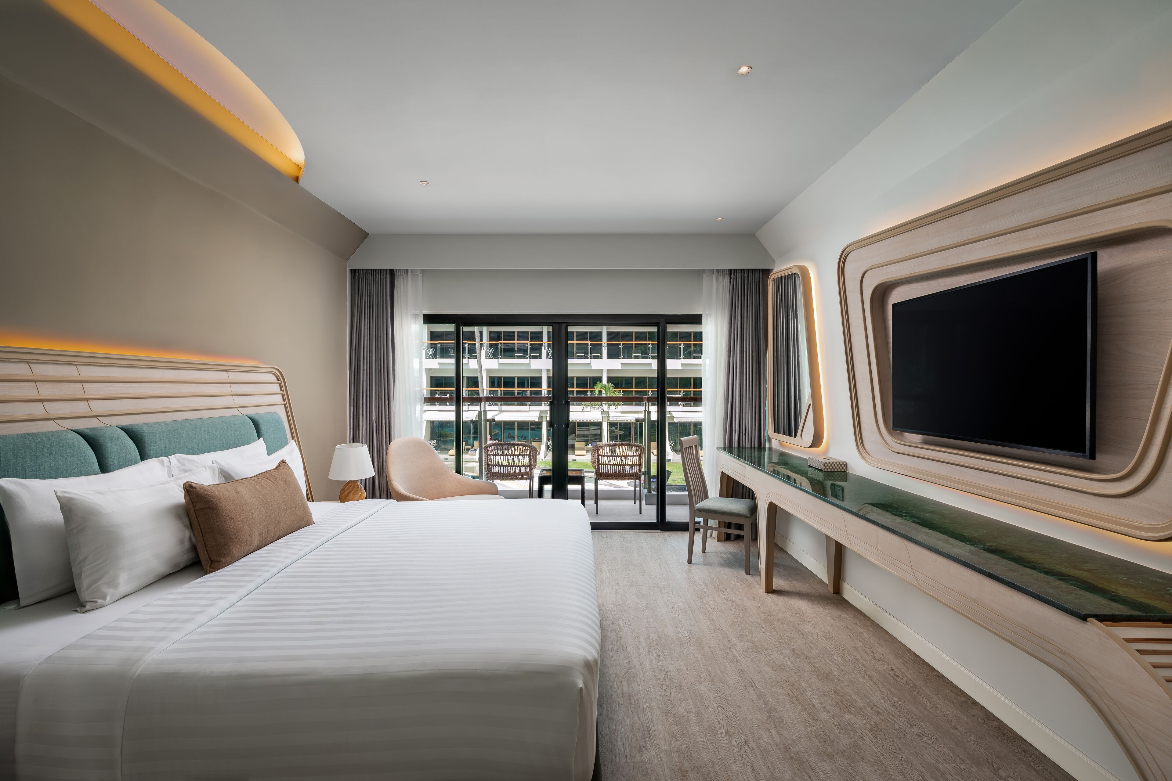 A hotel room with a large bed, a flat-screen TV mounted on the wall, a desk with a chair, and a view of the outside through large windows.