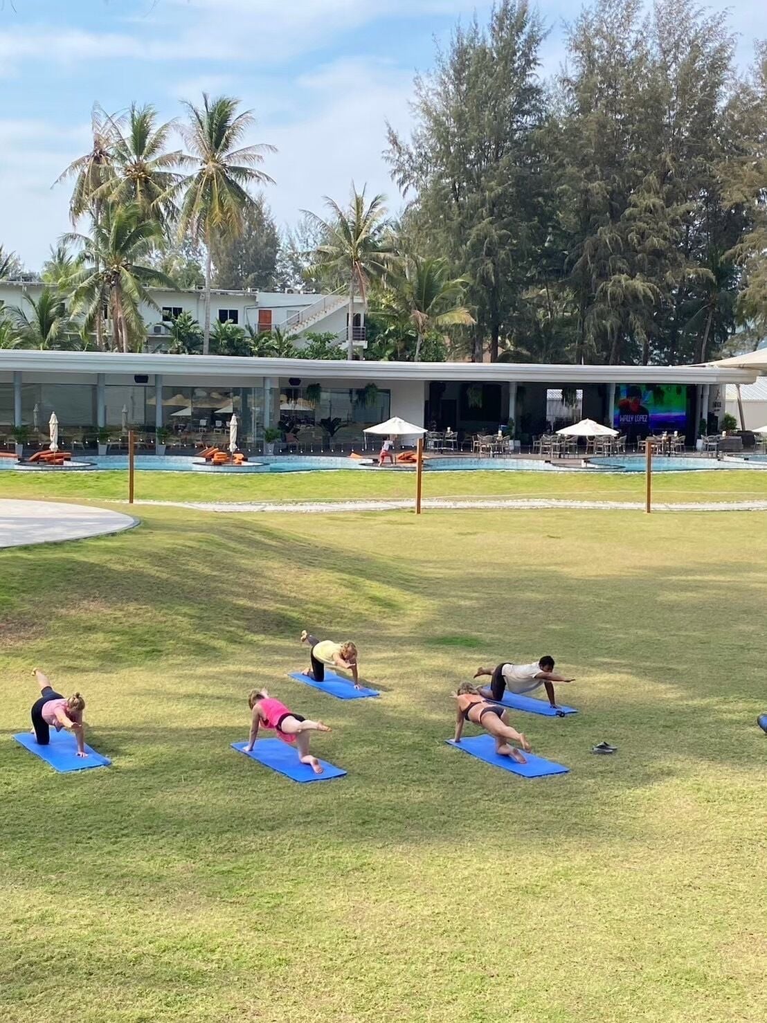 Eine Gruppe von Menschen praktiziert Yoga auf blauen Matten in einer grünen Freifläche. Im Hintergrund ist ein Pool und Liegestühle zu sehen.