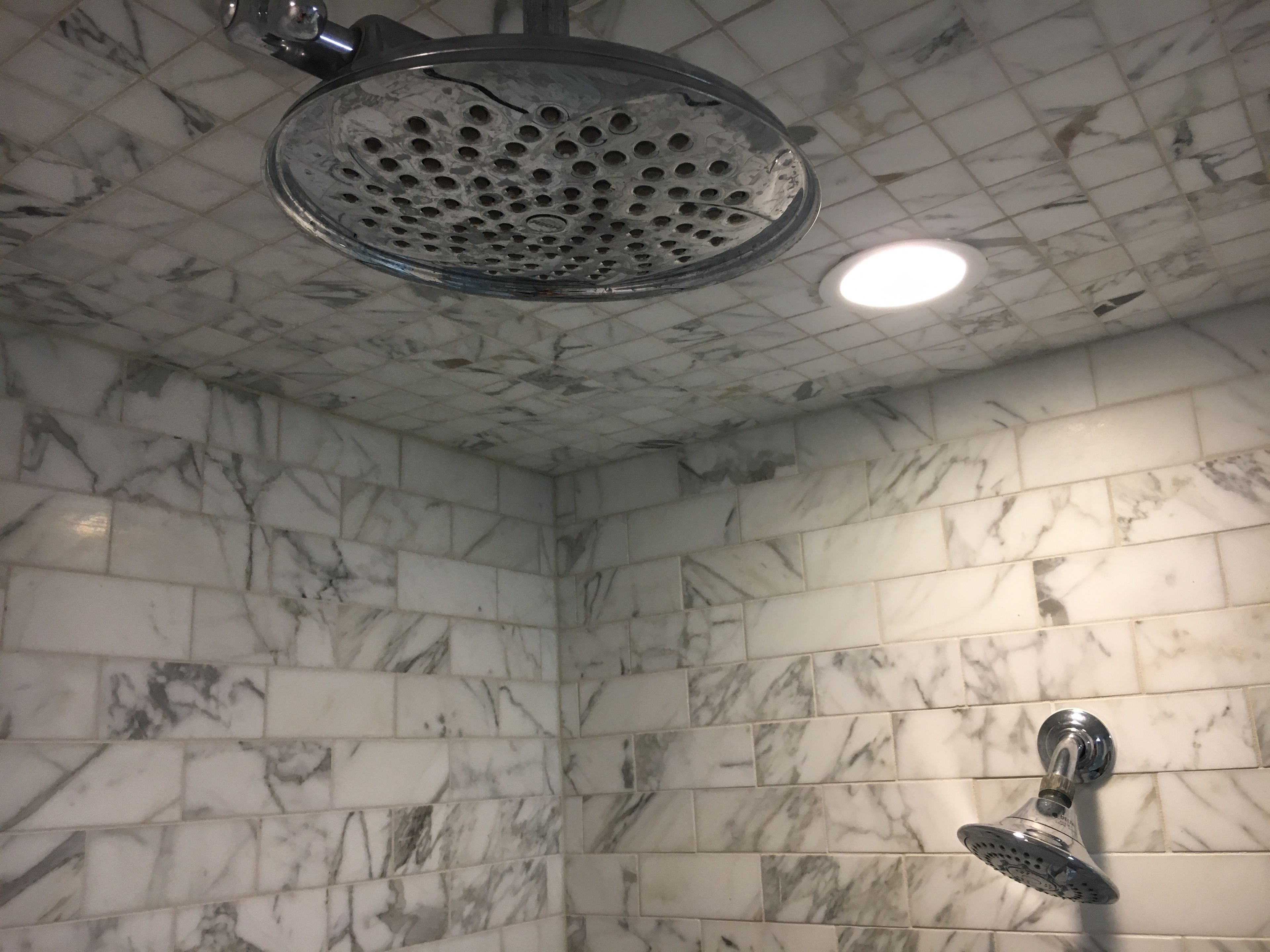 A shower with a rain showerhead and a handheld showerhead, set in a room with marble-patterned tiled walls.