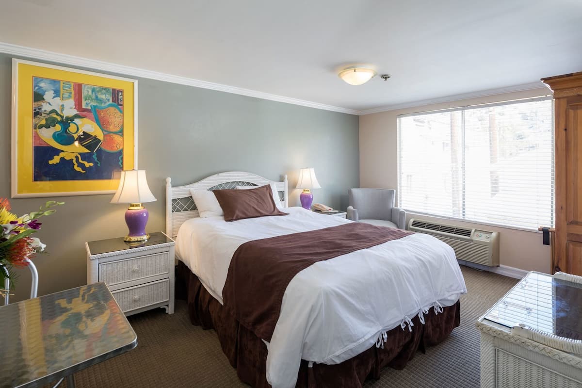 Quarto de hotel com uma cama, duas mesas de cabeceira, uma cadeira, janela com persianas e um quadro na parede.