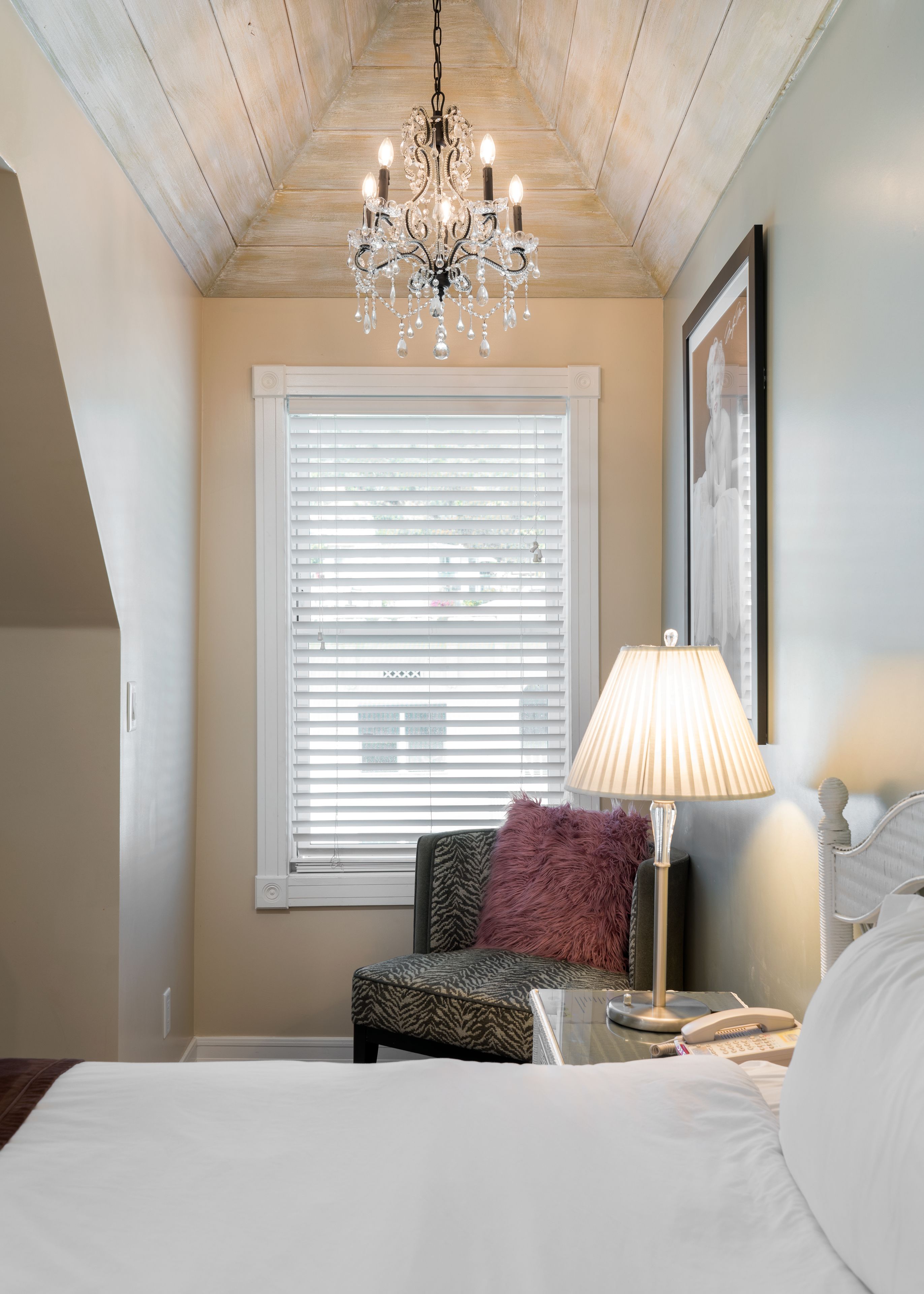 Une chambre avec un grand lit, un lustre, un fauteuil agrémenté d’un coussin moelleux, une table de chevet avec une lampe et une fenêtre avec des stores.