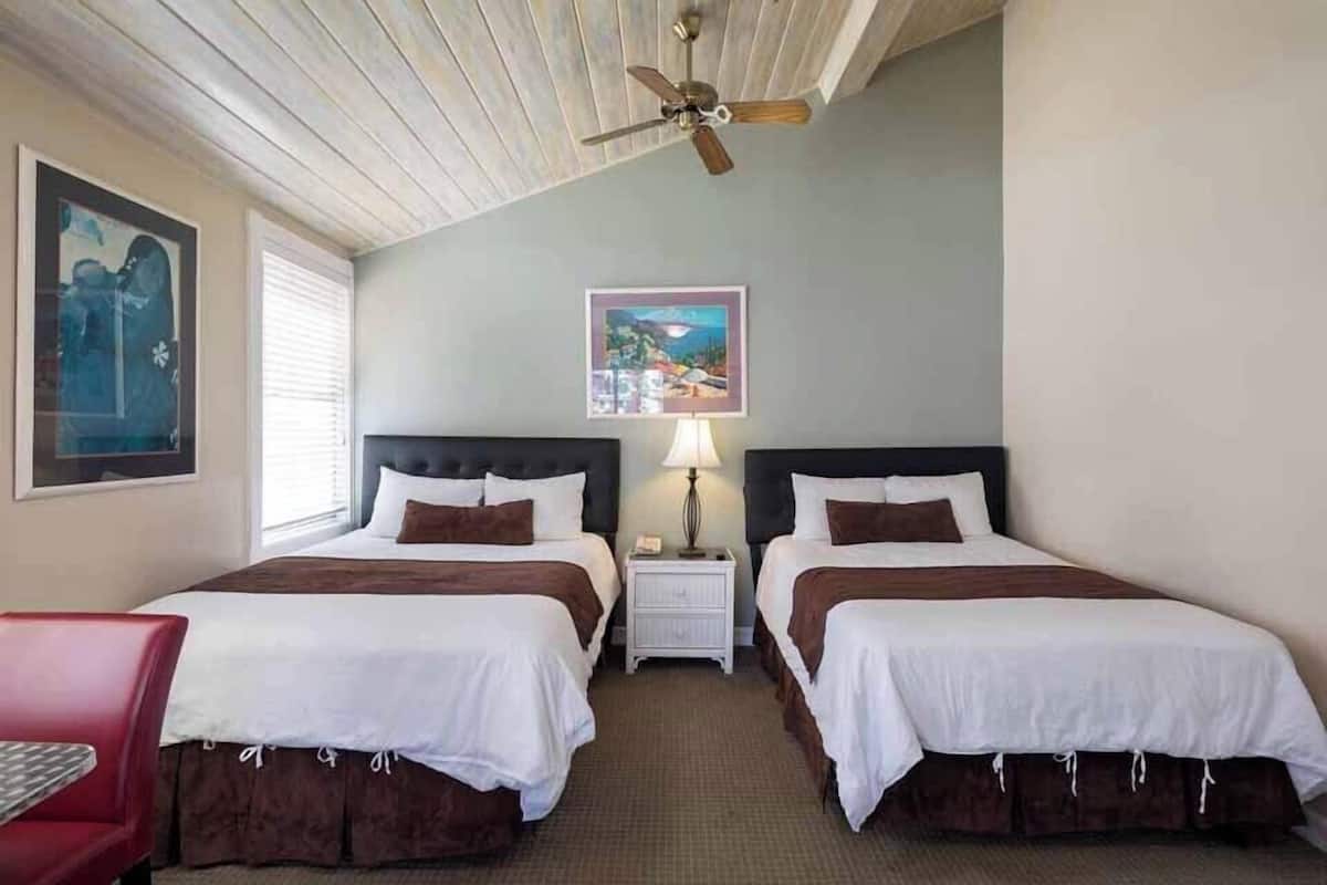 Quarto de hotel com duas camas, um ventilador de teto, mesa de cabeceira com luminária e quadros nas paredes.