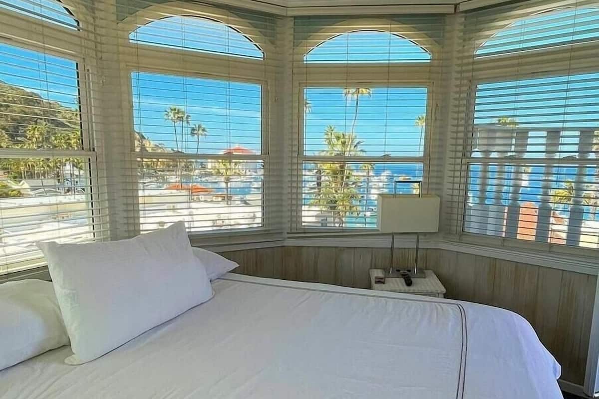 Quarto de hotel com uma cama grande, uma mesinha de cabeceira e um ventilador de teto. Há janelas amplas com vista para a praia e palmeiras.