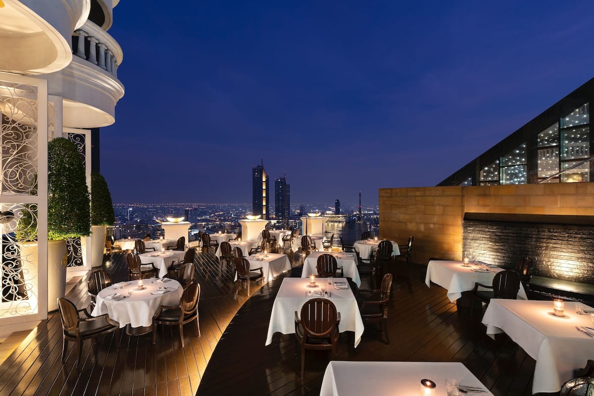 A rooftop restaurant with tables and chairs, a city view, and a modern building in the background.