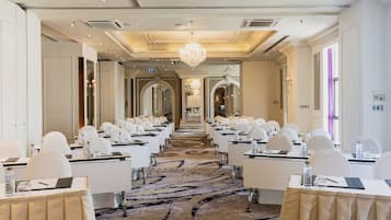 Un salón de conferencias con filas de mesas y sillas, alfombra estampada y un candelabro.