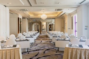 Un salón de conferencias con filas de mesas y sillas, alfombra estampada y un candelabro.