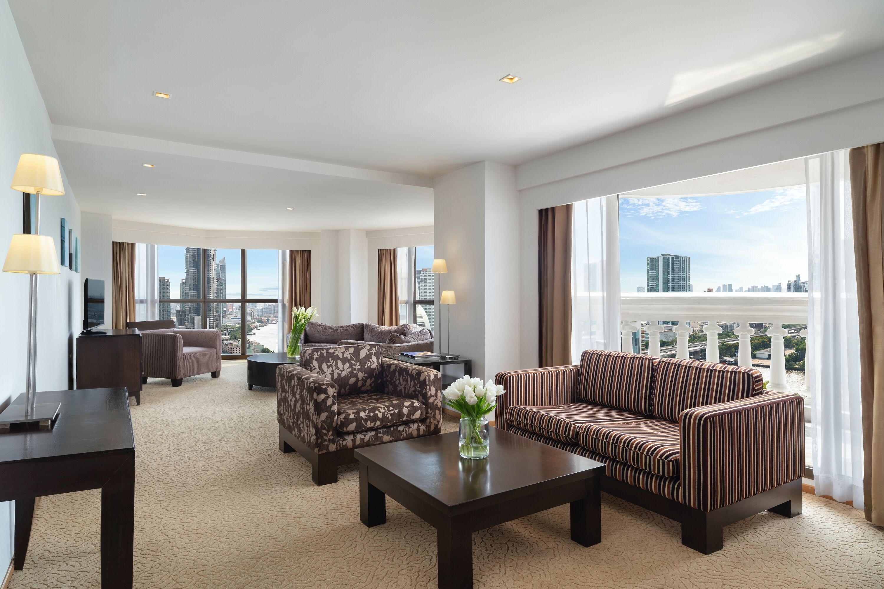 A spacious hotel room with a large window offering a city view, a sofa, a chair, and a coffee table.