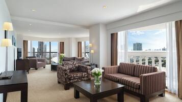 A spacious hotel room with a large window offering a city view, a sofa, a chair, and a coffee table.
