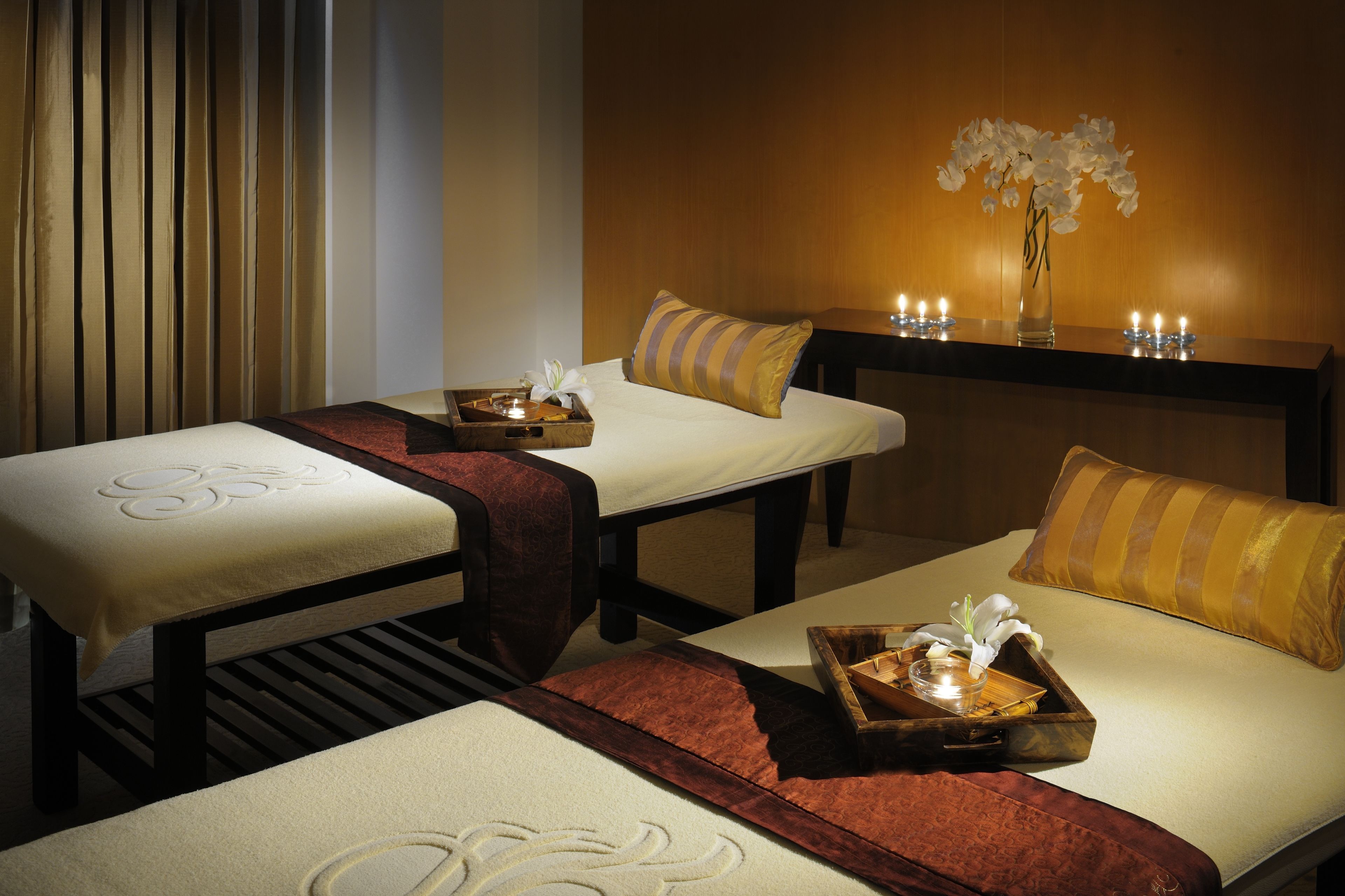 A spa room with two massage tables, each with a tray and candle, and a vase with white flowers on the wall.